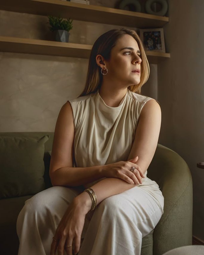 Jamille Façanha, de blusa e calça off-white, sentada num sofá verde-oliva, olhando pela janela sob uma luz lateral quente.